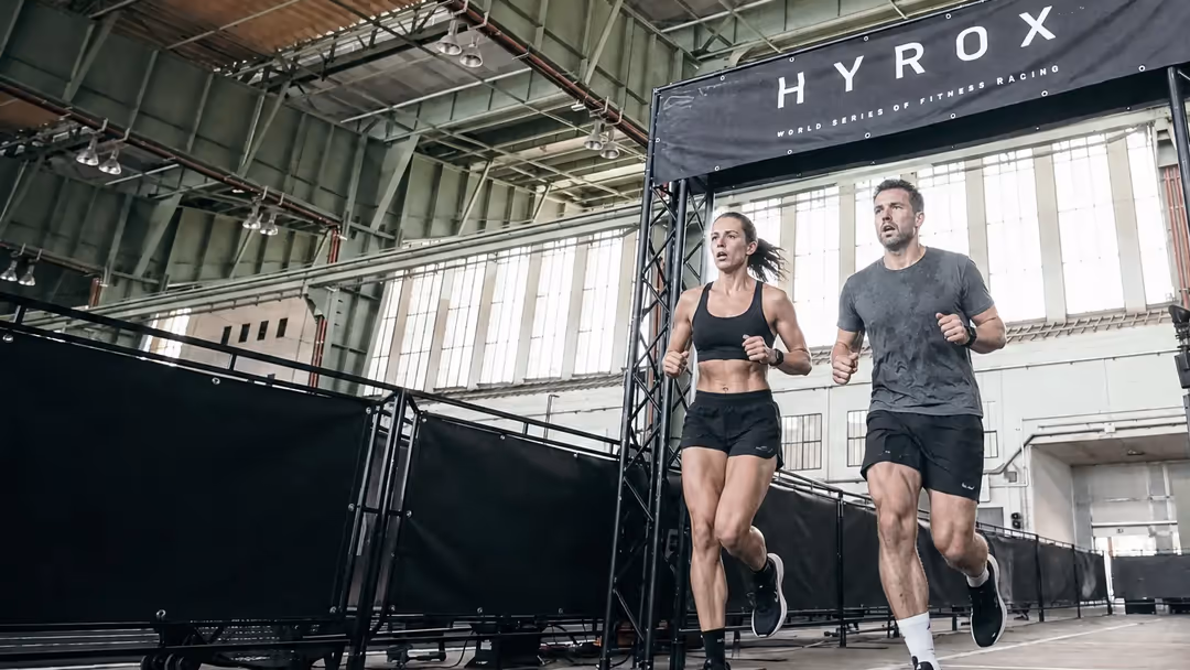 Zwei Läufer im HYROX-Rennen, unter dem HYROX-Banner in der Halle