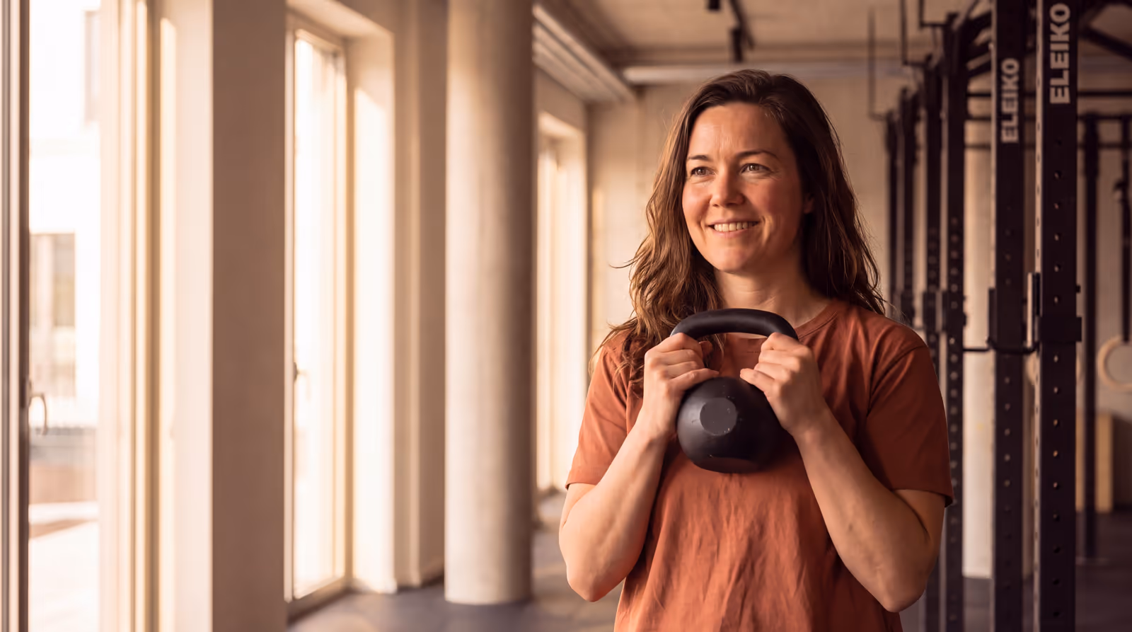Eine Frau hält lächelnd eine Kettlebell vor der Brust, im Studio am Fenster